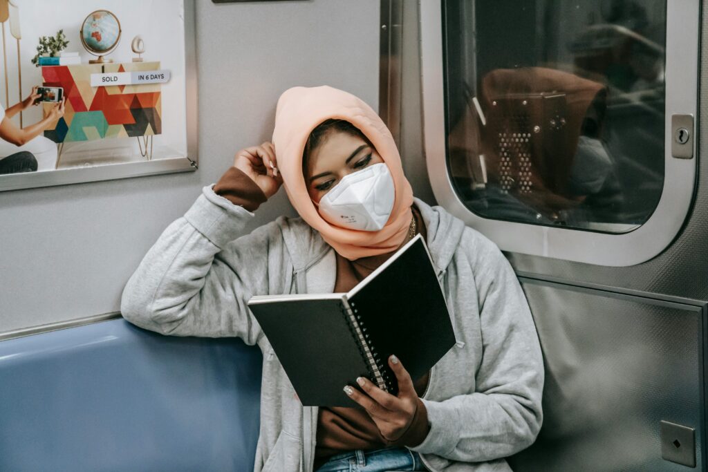 young Muslim ethnic woman in casual outfit and hijab with protective mask reading notebook while sitting in train in metro